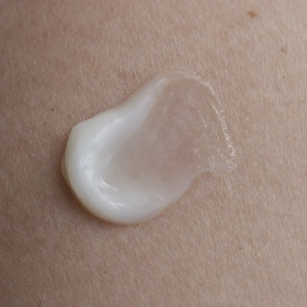 Extreme close-up of a small amount of pale cream applied to skin, its edges thinning into a nearly transparent film against the skin surface, photographed under even studio lighting.